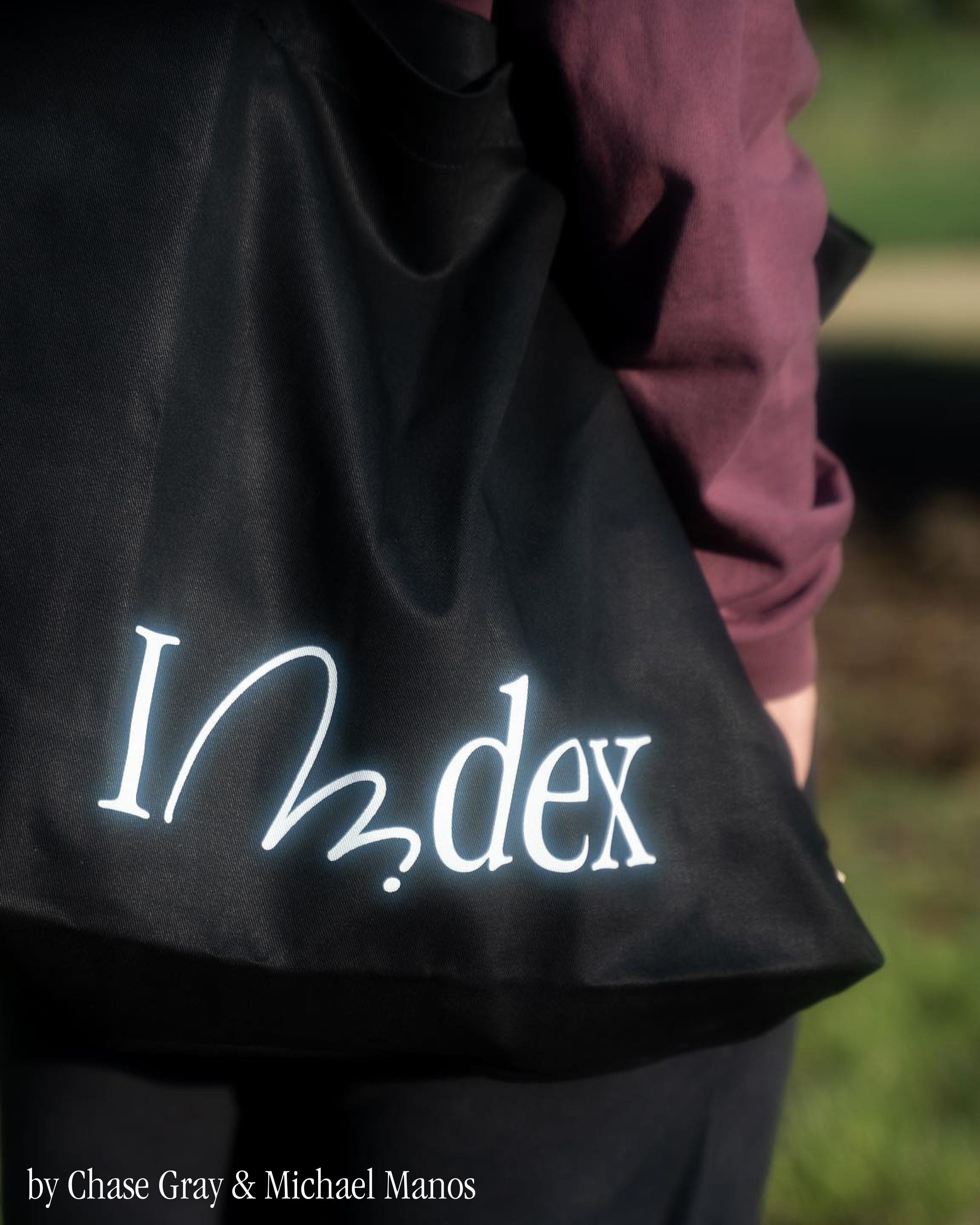 Black Index tote bag with the wordmark printed in white, carried on someone’s shoulder outdoors.