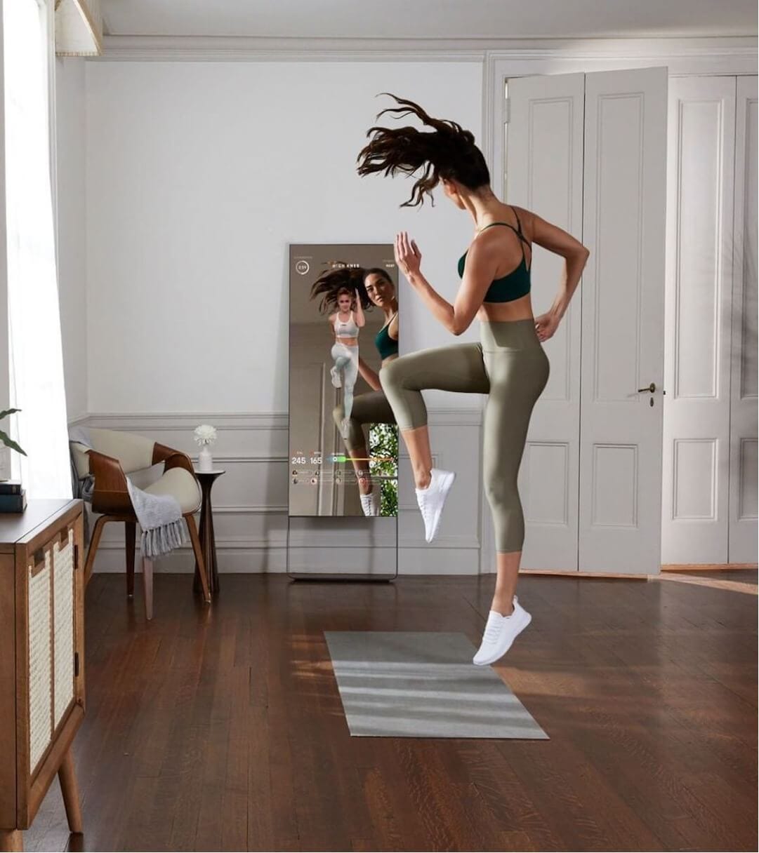 A person mid-jump in a living room workout in front of the Mirror device, which shows their reflection and live stats.