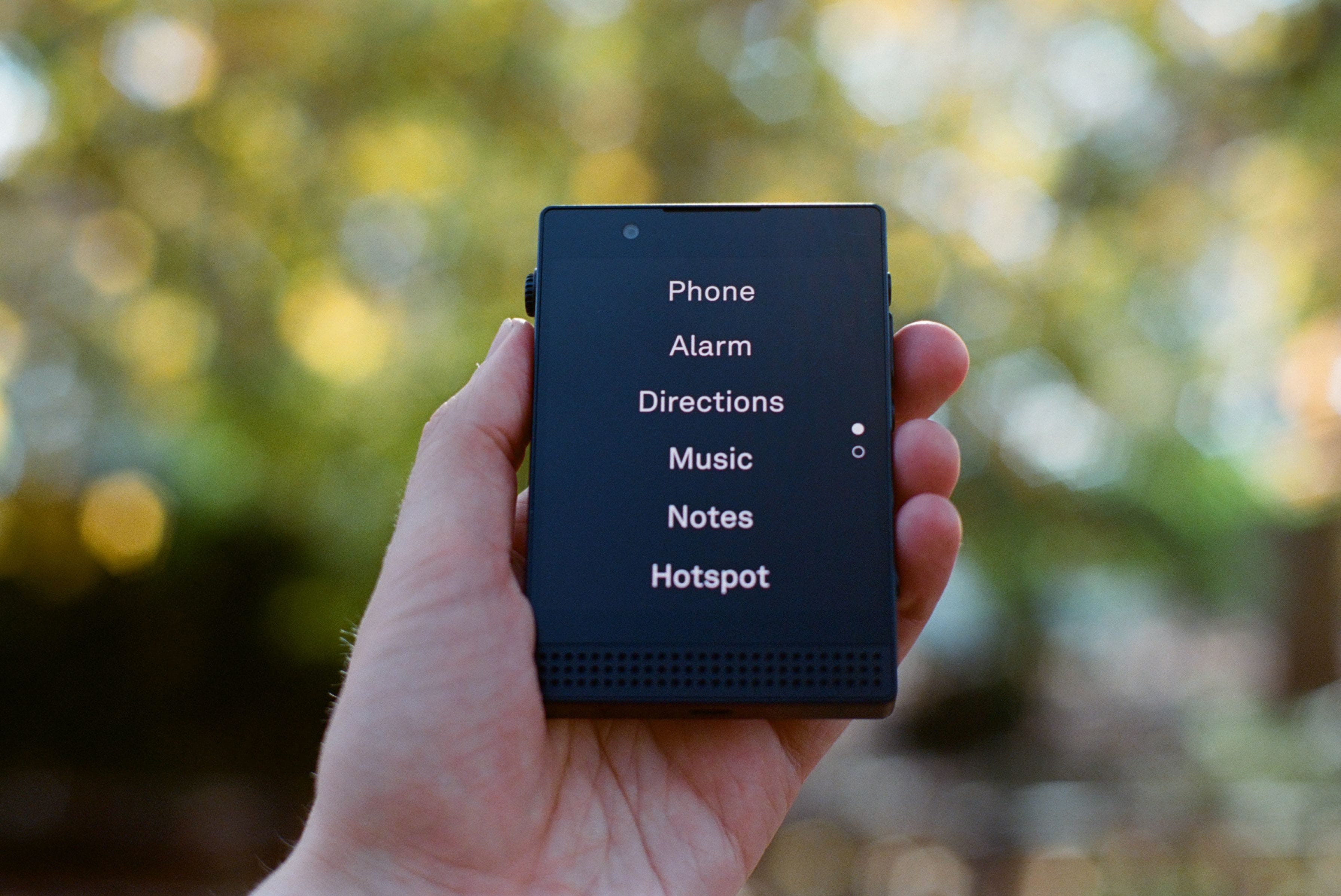 A hand holding the Light Phone III outdoors with the home menu visible on its EInk screen: Phone, Alarm, Directions, Music, Notes, Hotspot.