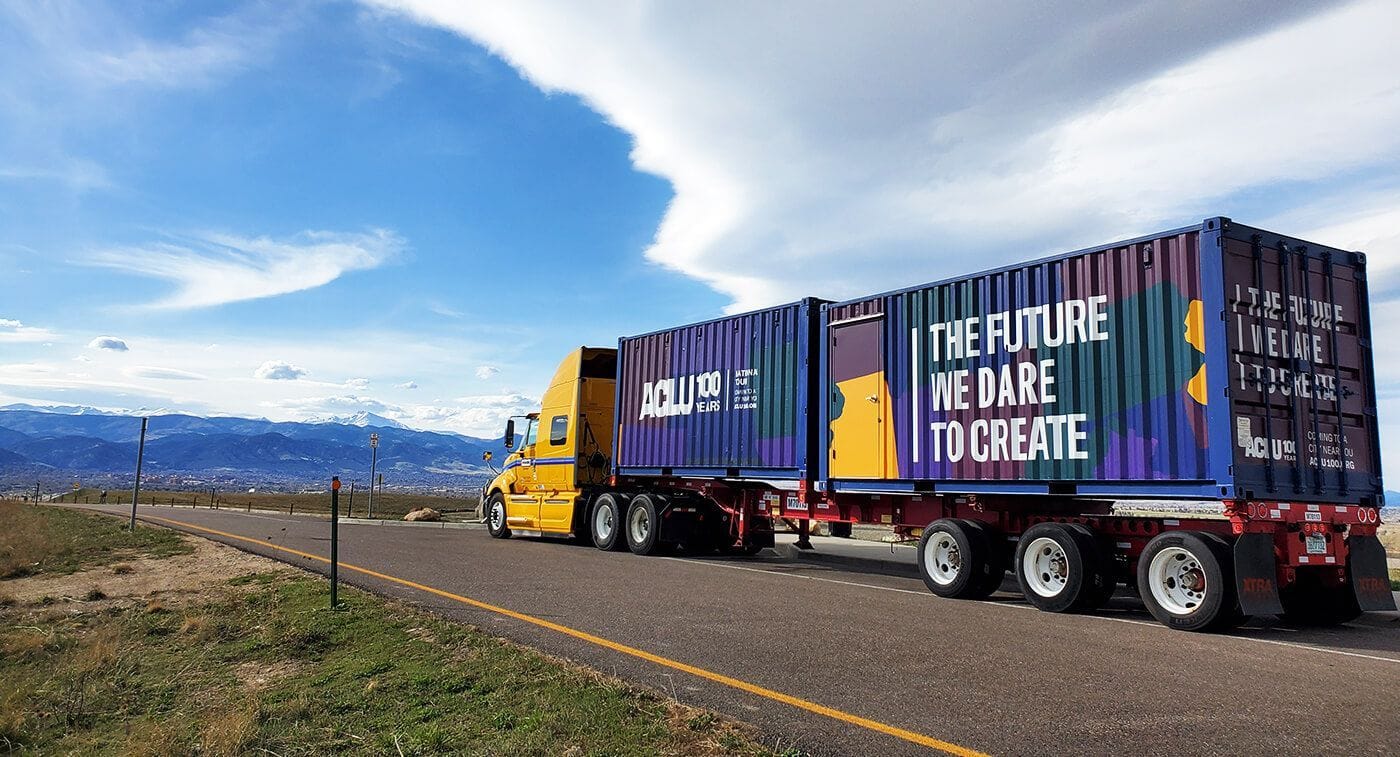 Yellow ACLU semi-truck pulling a multi-colored shipping container printed 'The future we dare to create', parked on a Western road with mountains behind.