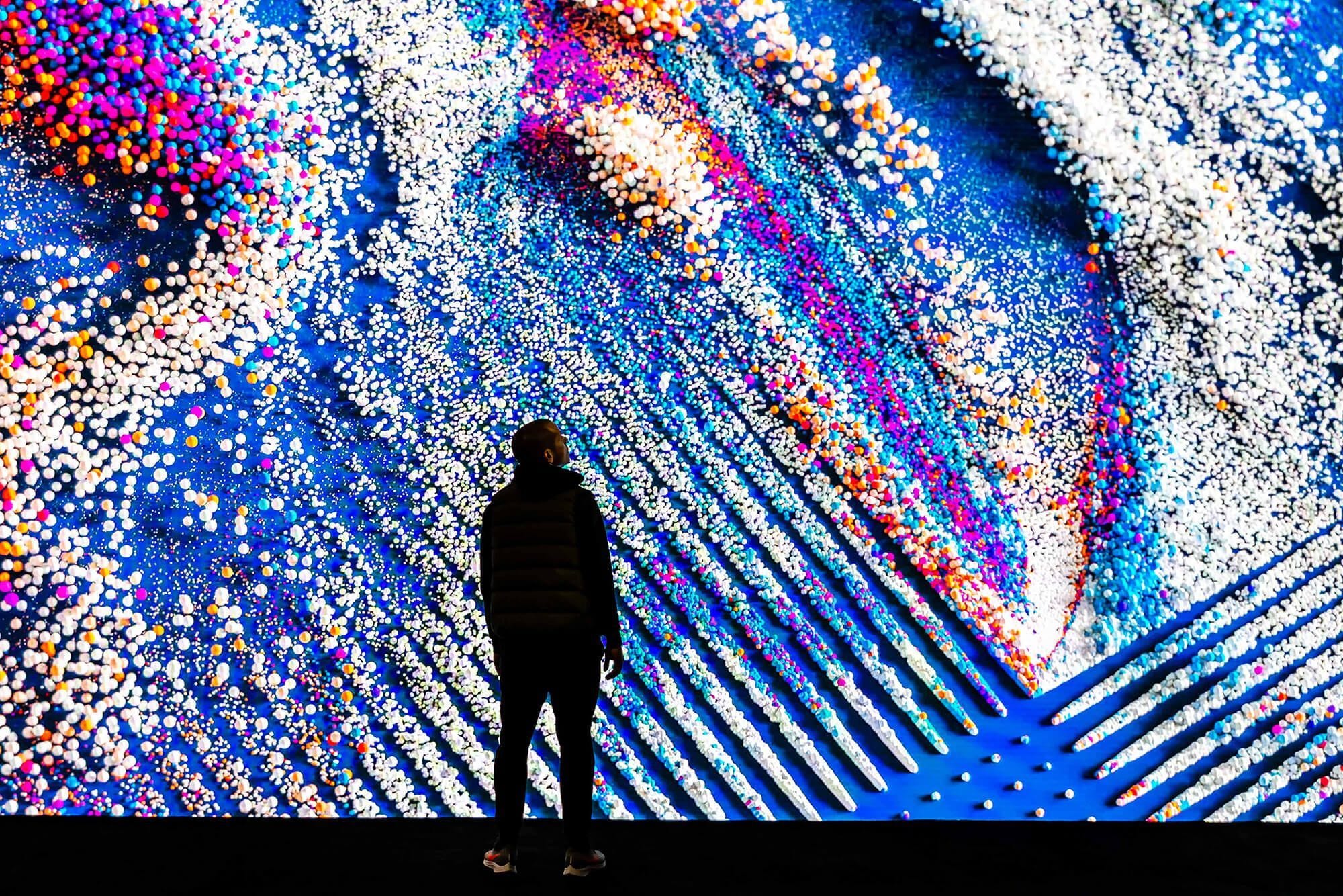 Photo of a man silhouetted in front of a large screen showing an IBM Think brand video of colorful particles.