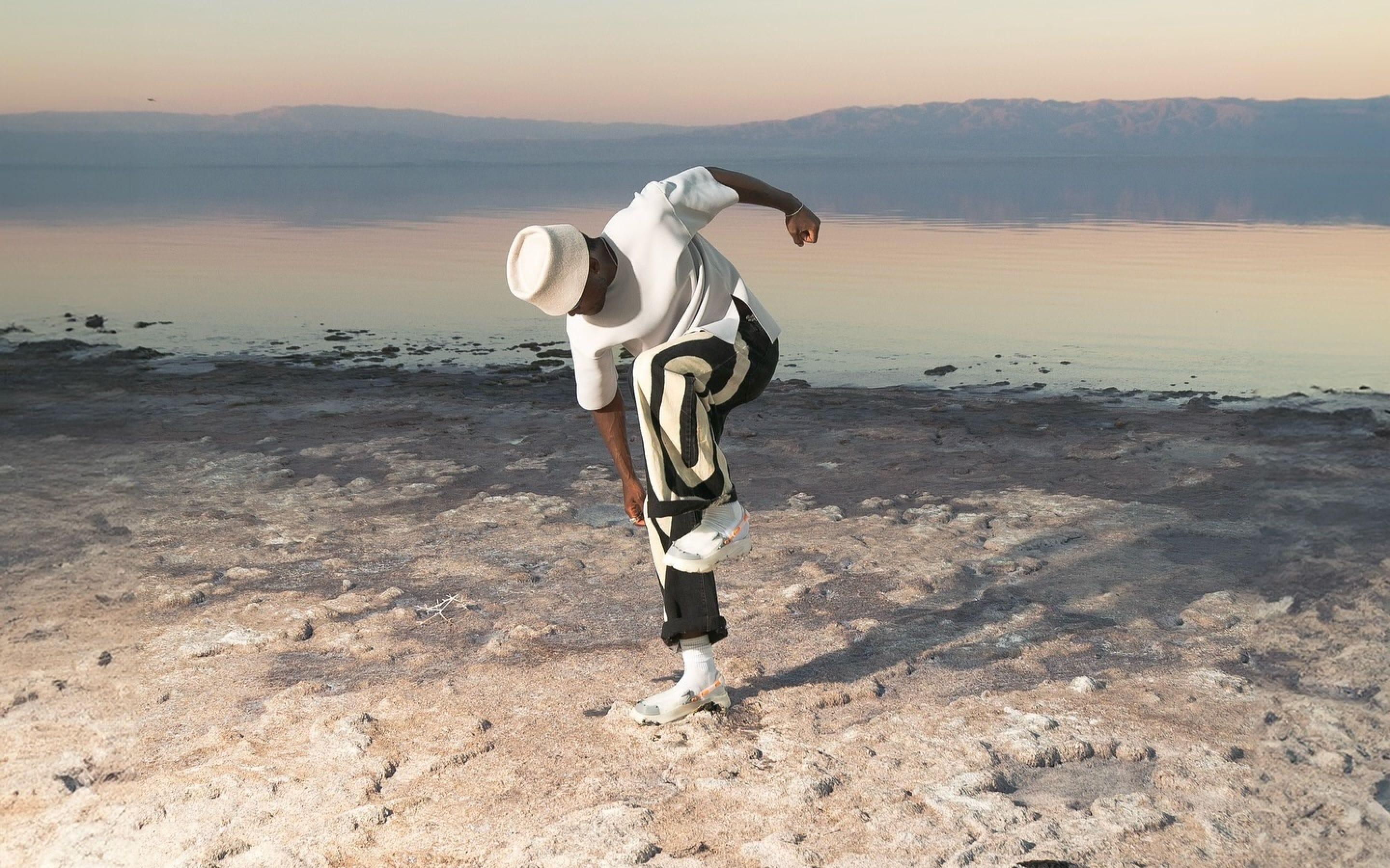 A model bent forward at a salt flat shoreline, wearing Presq garments and sneakers.