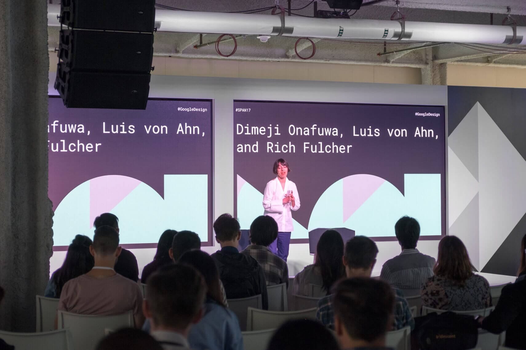 Photo of stage with a woman giving a speech and Google SPAN shapes on displays.
